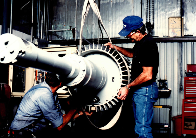 Fitting the generator rotor to the main shaft in the workshop — the finned rotor slides onto the shaft taper, sling-supported until the fit is drawn up. The same shaft carries the rotor hub at its far end.