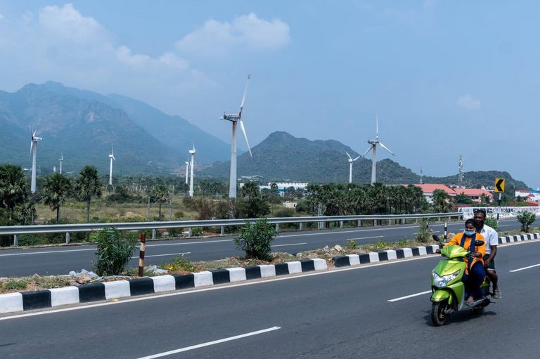 Scooter riders on a highway with rows of wind turbines and mountains behind near Muppandal, Tamil Nadu