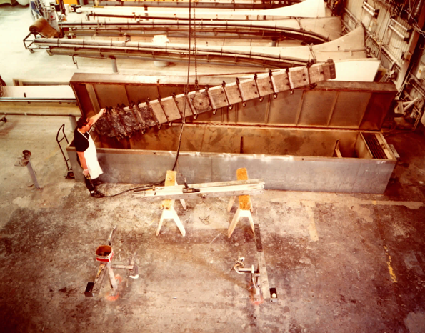 Blade spars over the treatment tank in the Carter factory — each welded steel spar is hot-dip protected end-to-end before the aerofoil shell goes on. Finished spars queue on the racks behind.