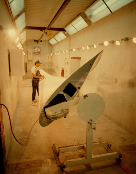 Blade root and pitch hardware being finished in the paint shop — the blade sits in a rotating fixture so the surfacer and topcoat go on evenly around the full section.