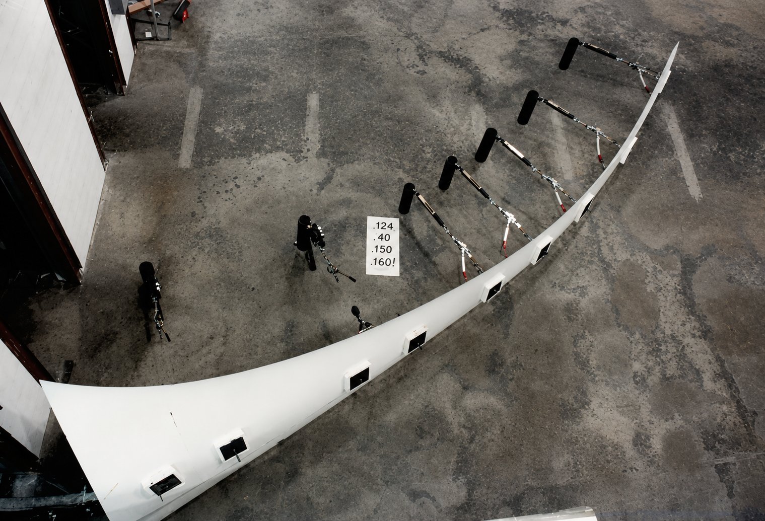 Aerial view of a full Carter rotor blade laid out on a workshop floor, instrumented with a row of loading actuators and weights along its length for a full-scale bending test