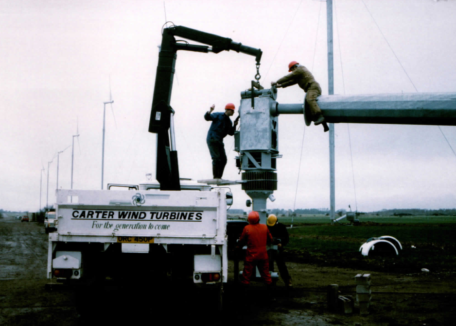 Fitting the yaw head and drivetrain stack straight from the service truck — ‘Carter Wind Turbines: for the generation to come’. The truck’s own crane is the only lift on site; the row of completed machines at Great Orton stands in the background.